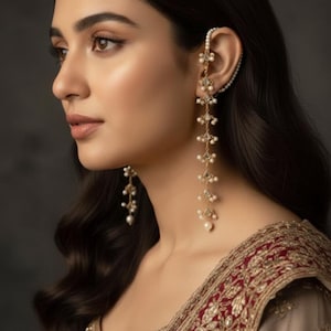 May include: An elegant gold ear cuff and dangling earrings with pearl details. The earrings cascade downwards. The woman is wearing a red and gold embroidered garment. The overall style is sophisticated and ornate.