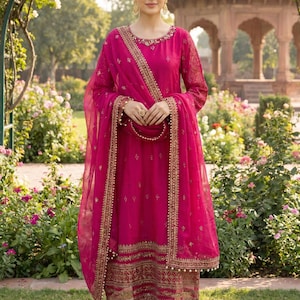 Puede incluir: Un conjunto salwar kameez magenta de manga larga con bordados dorados y un dupatta a juego. El atuendo presenta detalles intrincados alrededor del escote, las mangas y el dobladillo. El modelo está en un jardín.