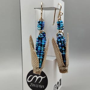 May include: A pair of blue and white beaded corn cob earrings with gold hooks. The earrings are displayed on a white stand with the logo "cm Zinapsys".