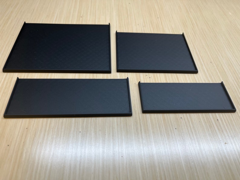 Peut inclure: Quatre plateaux rectangulaires noirs de tailles diff&eacute;rentes, dispos&eacute;s en rang&eacute;e sur une surface en bois clair.