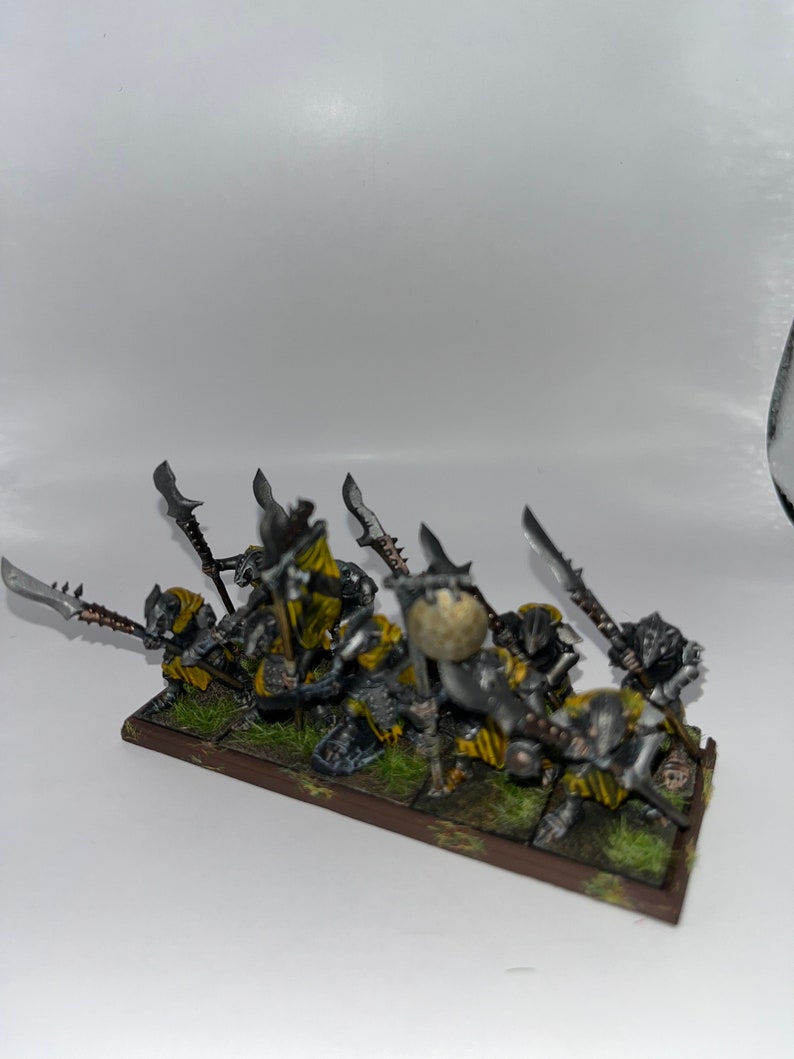 Peut inclure: Un groupe de huit figurines miniatures peintes en gris et jaune tenant des lances et des boucliers. Les figurines sont debout sur une base brune avec de l'herbe verte.