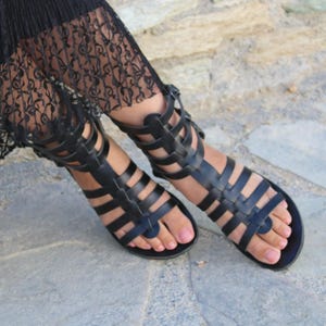 Puede incluir: Sandalias gladiadoras de cuero negro con múltiples correas y una presilla para el dedo gordo. Las sandalias tienen suela plana y se usan sobre una superficie de piedra. Las sandalias son una opción de calzado elegante.
