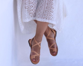 Può includere: Sandali in pelle color cuoio con più cinturini e un anello per le dita. I sandali hanno una suola piatta. Le calzature sono mostrate su uno sfondo bianco, suggerendo un focus sui sandali.