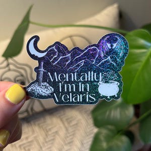 以下が含まれることがあります： 三日月、剣、山脈、煙の出ている釜、そして「Mentally I'm in Velaris」というテキストが描かれた、黒と白のデザインのホログラフィックステッカーです。