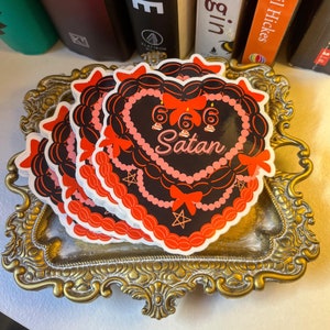 May include: Three black and red stickers with a heart-shaped cake design. The cake is decorated with red bows, black beads, and white stars. The cake has the numbers 666 and the word "Satan" written on it.