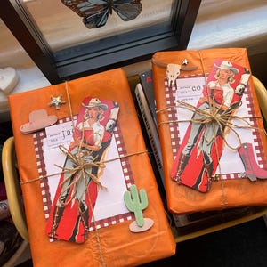 May include: Two books wrapped in orange paper with a red and white checkered pattern. The books are decorated with cowboy-themed paper cutouts, including a woman in a cowboy hat and boots, a cactus, and a cowboy hat. The books are tied with twine.