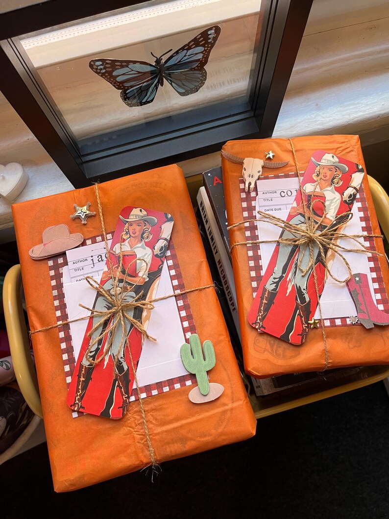 May include: Two books wrapped in orange paper with a red and white checkered pattern. The books are decorated with cowboy-themed paper cutouts, including a woman in a cowboy hat and boots, a cactus, and a cowboy hat. The books are tied with twine.