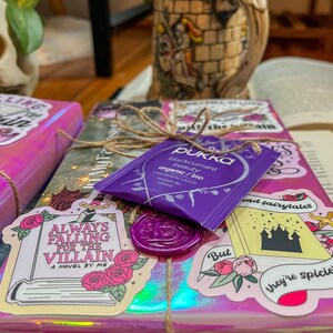 K&ouml;nnte beinhalten: Ein pink- und lilafarbenes, verpacktes Buch mit einem lilafarbenen Wachssiegel und einem Aufkleber mit der Aufschrift "Always Falling for the Villain, A Novel by Me". Es gibt auch einen Aufkleber mit der Aufschrift "But they're spicier" mit einer schwarzen Schlossillustration und einen Aufkleber mit der Aufschrift "Read Fairytales".