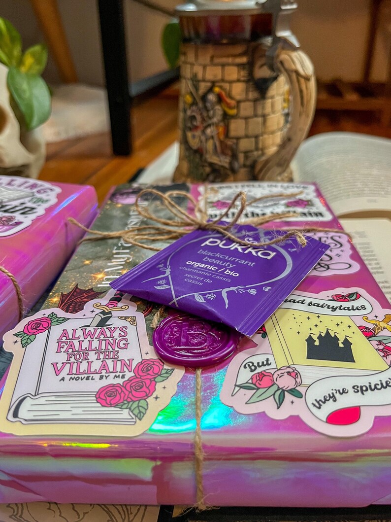 K&ouml;nnte beinhalten: Ein pink- und lilafarbenes, verpacktes Buch mit einem lilafarbenen Wachssiegel und einem Aufkleber mit der Aufschrift "Always Falling for the Villain, A Novel by Me". Es gibt auch einen Aufkleber mit der Aufschrift "But they're spicier" mit einer schwarzen Schlossillustration und einen Aufkleber mit der Aufschrift "Read Fairytales".