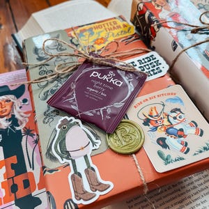 May include: A wrapped gift box with a brown paper cover and a green wax seal. The box is decorated with stickers of a frog wearing cowboy boots, a horse and rider, and a tea bag with the text "Pukka night time berry organic/bio" on it. The box is tied with twine and has a sticker that says "Ride or Die Bitches".