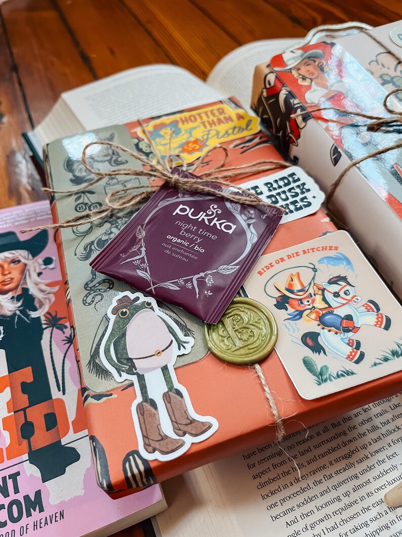 May include: A wrapped gift box with a brown paper cover and a green wax seal. The box is decorated with stickers of a frog wearing cowboy boots, a horse and rider, and a tea bag with the text "Pukka night time berry organic/bio" on it. The box is tied with twine and has a sticker that says "Ride or Die Bitches".