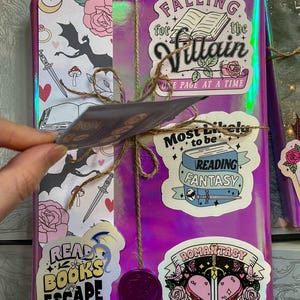K&ouml;nnte beinhalten: Ein in lila Geschenkpapier verpacktes Buch mit verschiedenen Aufklebern. Die Aufkleber zeigen einen Drachen, ein Buch mit dem Text "Falling for the Villain, One Page at a Time", ein Buch mit dem Text "Most Likely to be Reading Fantasy" und ein Buch mit dem Text "Romanstasy Book Club".