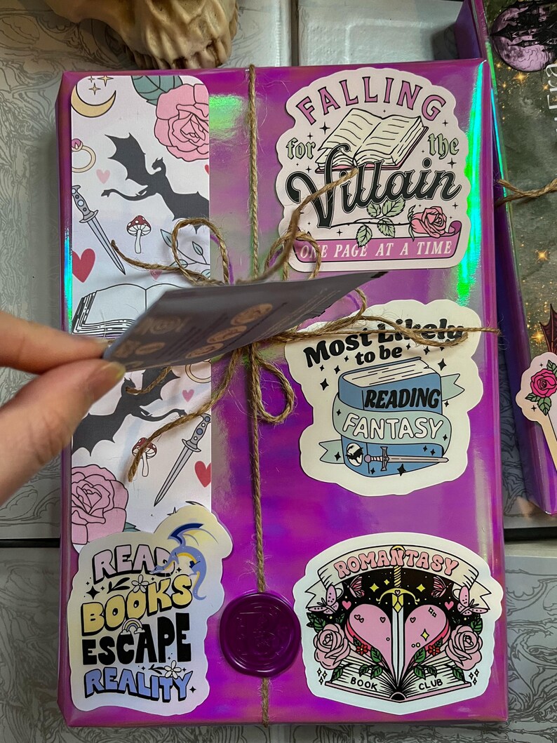 K&ouml;nnte beinhalten: Ein in lila Geschenkpapier verpacktes Buch mit verschiedenen Aufklebern. Die Aufkleber zeigen einen Drachen, ein Buch mit dem Text "Falling for the Villain, One Page at a Time", ein Buch mit dem Text "Most Likely to be Reading Fantasy" und ein Buch mit dem Text "Romanstasy Book Club".