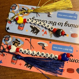 May include: Two beaded keychains with tassels. One keychain spells "ROZANOV" with a yellow heart and a black heart bead. The other keychain spells "HOLLANDER" with a white heart and a blue heart bead.