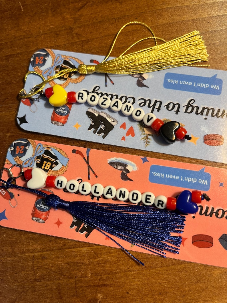 May include: Two beaded keychains with tassels. One keychain spells "ROZANOV" with a yellow heart and a black heart bead. The other keychain spells "HOLLANDER" with a white heart and a blue heart bead.