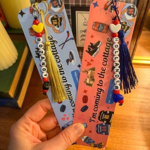 May include: Two decorative bookmarks with beaded charms and tassels. One is light blue with the name "ROZANOV" and the text "coming to the cottage". The other is pink with the name "HOLLANDER" and the text "I'm coming to the cottage".