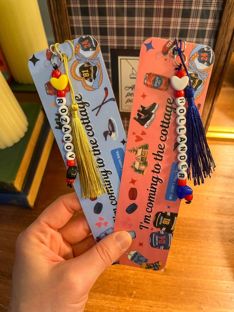 May include: Two decorative bookmarks with beaded charms and tassels. One is light blue with the name "ROZANOV" and the text "coming to the cottage". The other is pink with the name "HOLLANDER" and the text "I'm coming to the cottage".