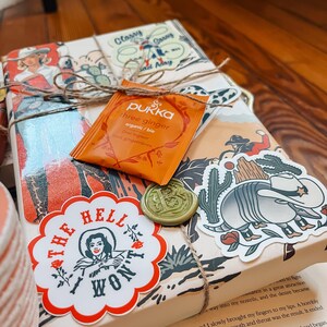 May include: A wrapped book with a brown paper cover and a twine bow. The book has a sticker with the text "The Hell I Won't" and a sticker of a cartoon armadillo. There is a small orange tea bag with the text "Pukka Three Ginger Organic/Bio" on the front.