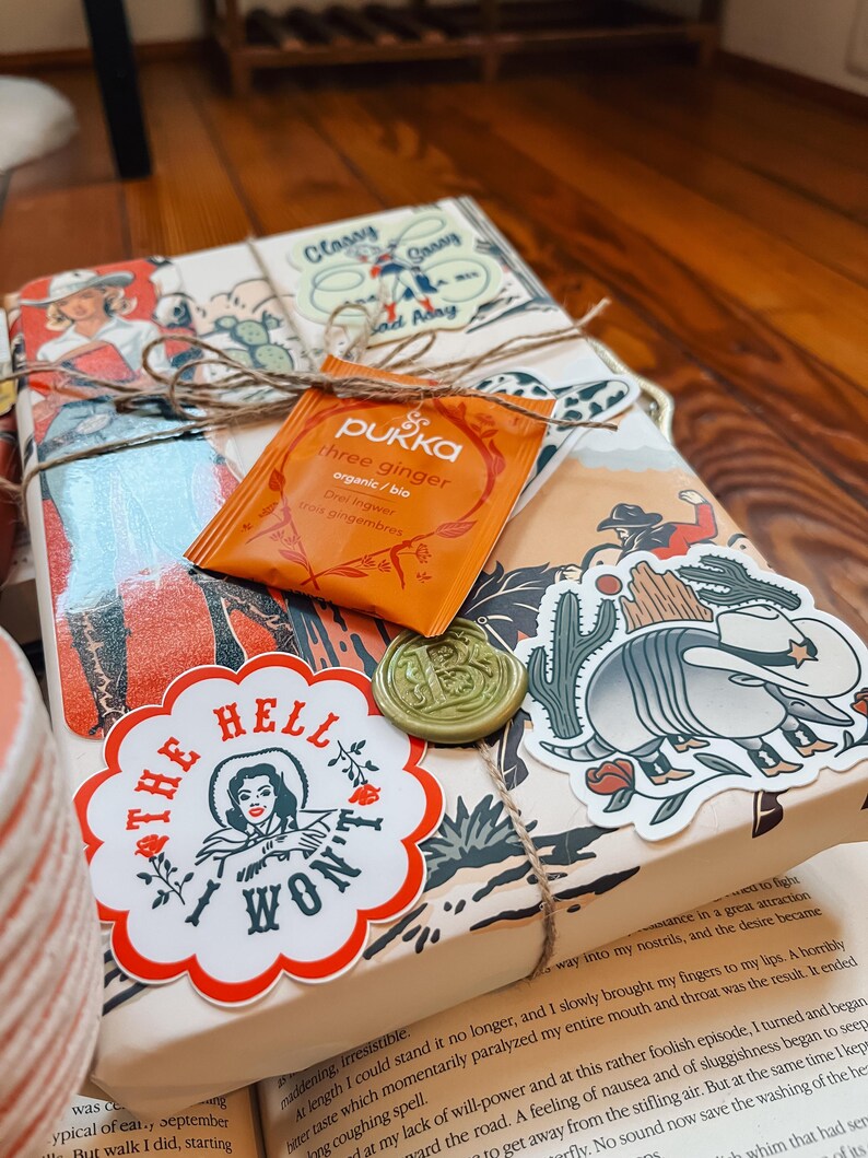 May include: A wrapped book with a brown paper cover and a twine bow. The book has a sticker with the text "The Hell I Won't" and a sticker of a cartoon armadillo. There is a small orange tea bag with the text "Pukka Three Ginger Organic/Bio" on the front.