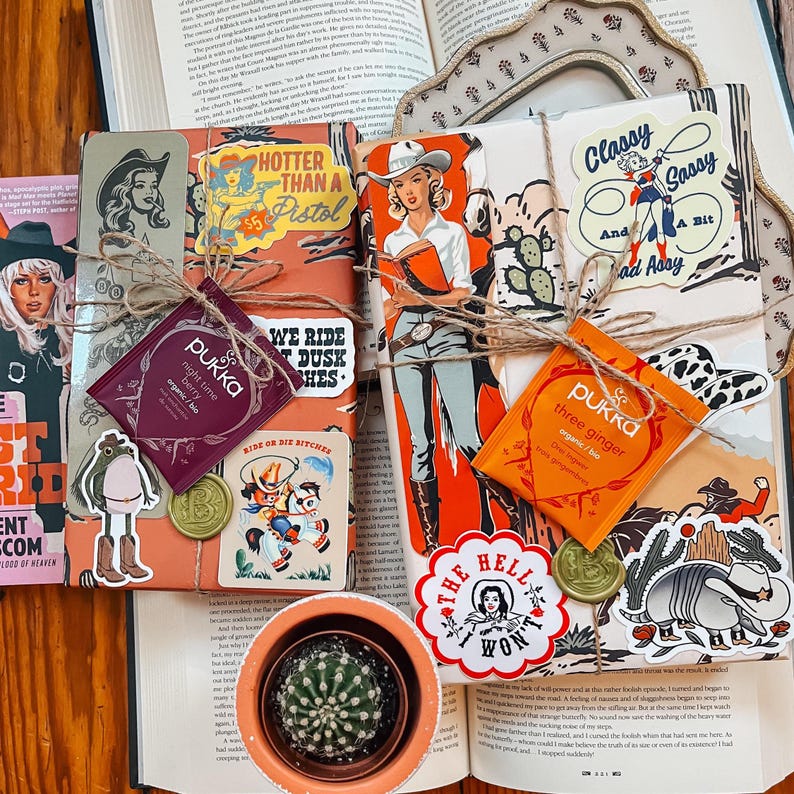 May include: A collection of wrapped books with a western theme. The books are decorated with stickers, twine, and tea bags. The stickers feature cowboys, horses, and sayings like "Hotter Than a Pistol" and "Ride or Die Bitches".