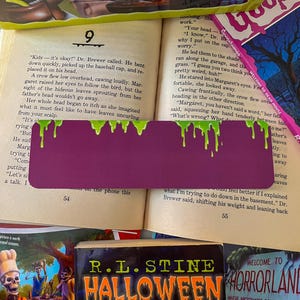 May include: A close-up of a purple bookmark with green dripping paint on it. The bookmark is placed on a page of a book titled "Goosebumps" with the text "Kids - it's okay!" and "What's wrong?" visible. The book is open to page 54.