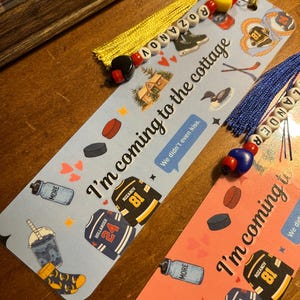May include: Two decorative bookmarks with tassels. One is light blue, the other is pink. Both have hockey-themed illustrations, including jerseys, hockey pucks, and the text "I'm coming to the cottage." One bookmark has the name "Rozanov" and the other "Hollander".