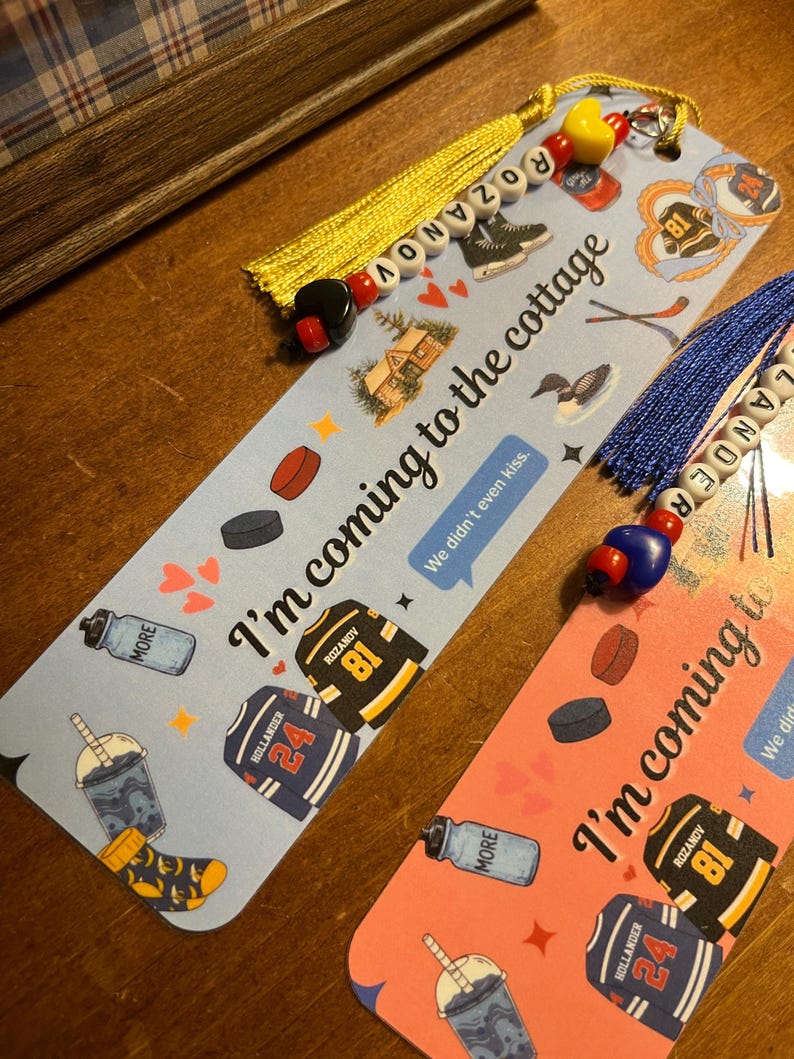May include: Two decorative bookmarks with tassels. One is light blue, the other is pink. Both have hockey-themed illustrations, including jerseys, hockey pucks, and the text "I'm coming to the cottage." One bookmark has the name "Rozanov" and the other "Hollander".