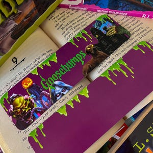 May include: A purple bookmark with green dripping slime. The bookmark features a Goosebumps logo and a scary monster with a mask. The text "Goosebumps" is printed on the bookmark. The bookmark is placed on top of a book with the text "Welcome to Dead House".