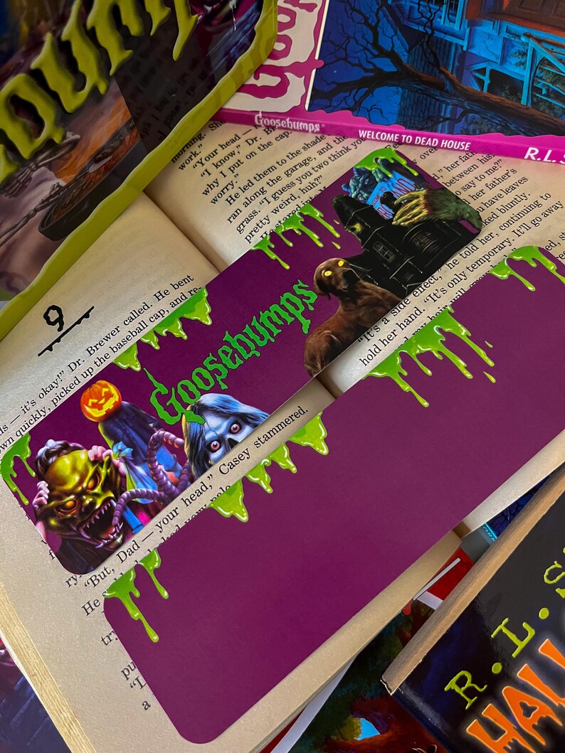 May include: A purple bookmark with green dripping slime. The bookmark features a Goosebumps logo and a scary monster with a mask. The text "Goosebumps" is printed on the bookmark. The bookmark is placed on top of a book with the text "Welcome to Dead House".