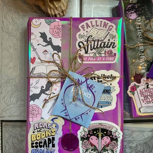 K&ouml;nnte beinhalten: Ein lila Geschenkpapier verpacktes Buch mit verschiedenen Aufklebern, darunter "Falling for the Villain", "Read Books Escape Reality", "Romantasy Book Club" und ein Teebeutel.