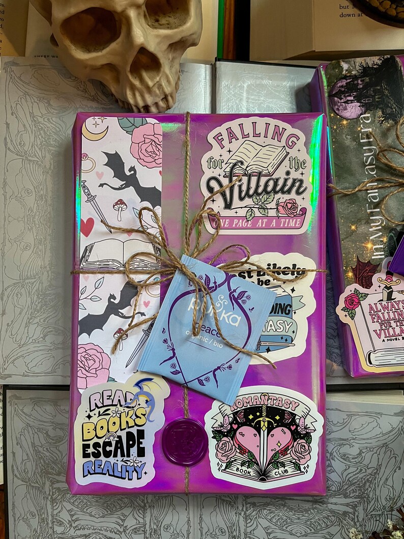 K&ouml;nnte beinhalten: Ein lila Geschenkpapier verpacktes Buch mit verschiedenen Aufklebern, darunter "Falling for the Villain", "Read Books Escape Reality", "Romantasy Book Club" und ein Teebeutel.