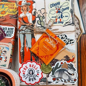 May include: A wrapped book with a variety of colorful stickers, including a sticker that says "Hotter Than a Pistol", a sticker that says "Classy Sassy and a Bit Bad Assy", and a sticker that says "The Hell I Won't". There is also a small orange tea bag with the text "pukka three ginger organic/bio Drei Ingwer trois gingembres".