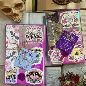 K&ouml;nnte beinhalten: Zwei verpackte Geschenke mit einem lila und pinken holografischen Papier. Die Geschenke sind mit Aufklebern und einem Teebeutel verziert. Die Aufkleber zeigen Designs zum Thema B&uuml;cher und Texte wie "Falling for the Villain", "Read Books Escape Reality" und "Always Falling for the Villain".