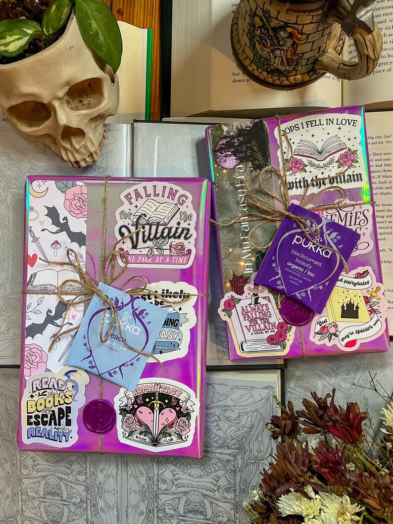 K&ouml;nnte beinhalten: Zwei verpackte Geschenke mit einem lila und pinken holografischen Papier. Die Geschenke sind mit Aufklebern und einem Teebeutel verziert. Die Aufkleber zeigen Designs zum Thema B&uuml;cher und Texte wie "Falling for the Villain", "Read Books Escape Reality" und "Always Falling for the Villain".