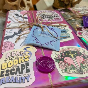 K&ouml;nnte beinhalten: Ein pinkfarbenes und schimmerndes Geschenkbuch mit einem violetten Siegel aus Wachs. Das Buch ist mit Aufklebern verziert, auf denen "Read Books Escape Reality", "Falling Villain", "Romanstasy" und "Peace" steht.