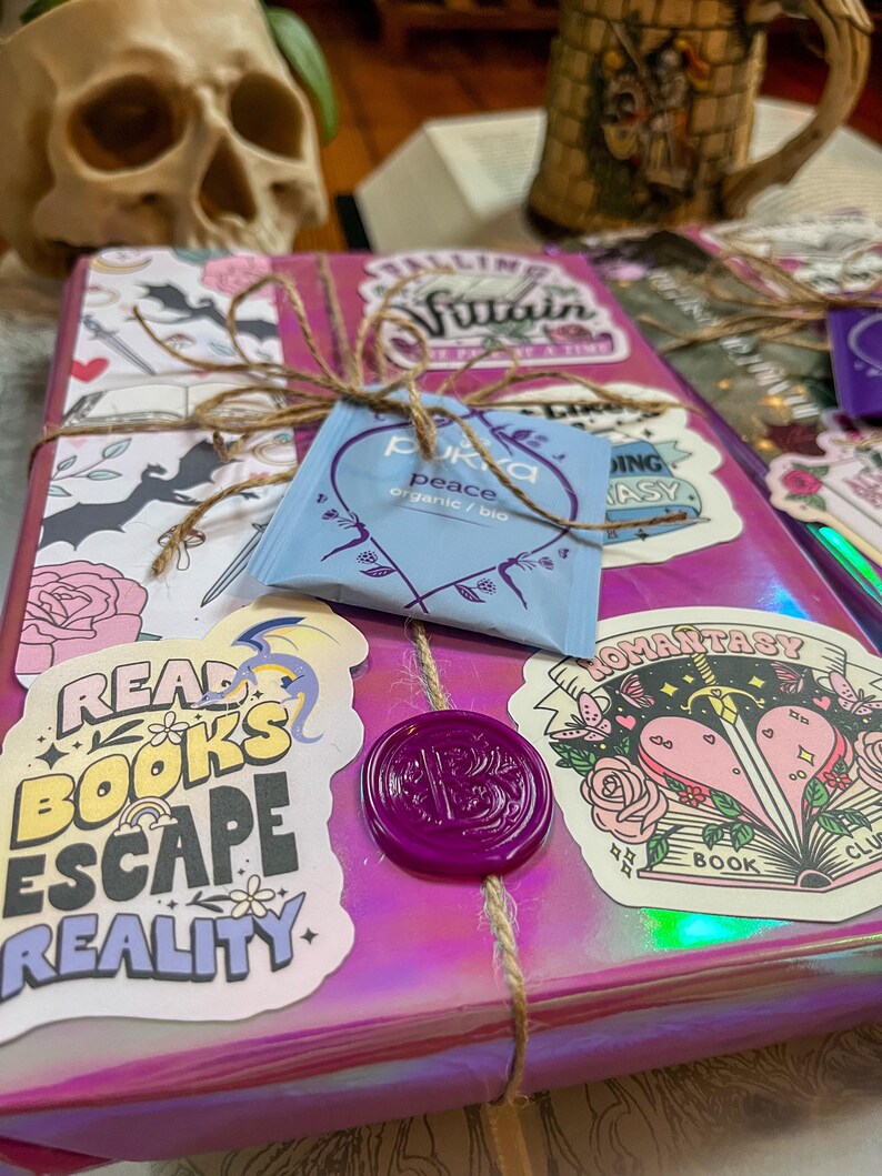 K&ouml;nnte beinhalten: Ein pinkfarbenes und schimmerndes Geschenkbuch mit einem violetten Siegel aus Wachs. Das Buch ist mit Aufklebern verziert, auf denen "Read Books Escape Reality", "Falling Villain", "Romanstasy" und "Peace" steht.
