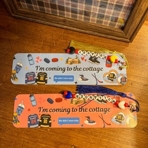 May include: Two decorative bookmarks with tassels. One is light blue, the other is coral. Both feature hockey-themed graphics, the text "I'm coming to the cottage", and the text "We didn't even kiss."