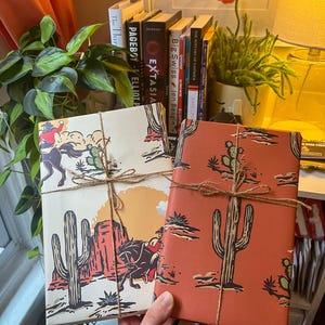 May include: Two wrapped gifts with a western desert theme. The gifts are wrapped in brown paper with a red and white cactus and cowboy design. The gifts are tied with twine.