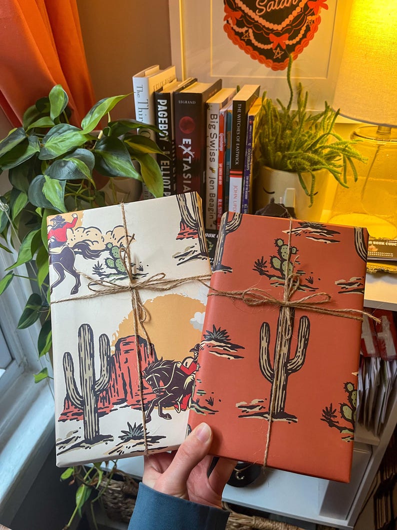 May include: Two wrapped gifts with a western desert theme. The gifts are wrapped in brown paper with a red and white cactus and cowboy design. The gifts are tied with twine.