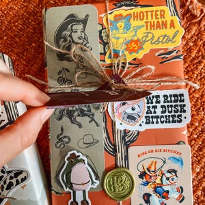 May include: A brown gift box wrapped in paper with a western theme. The paper features a cactus, a cowboy, and the text "Hotter than a Pistol". The box is decorated with several stickers, including a frog wearing cowboy boots, a woman riding a horse, and a seal with the text "Ride or Die Bitches".