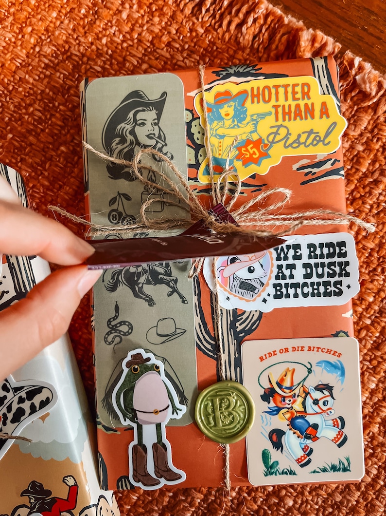 May include: A brown gift box wrapped in paper with a western theme. The paper features a cactus, a cowboy, and the text "Hotter than a Pistol". The box is decorated with several stickers, including a frog wearing cowboy boots, a woman riding a horse, and a seal with the text "Ride or Die Bitches".