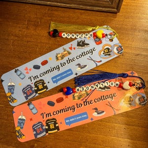 May include: Two decorative bookmarks with tassels and beaded accents. One is light blue, the other is coral. Both feature hockey-themed illustrations, text that reads "I'm coming to the cottage," and the names ROZANOV and HOLLANDER.