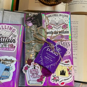 K&ouml;nnte beinhalten: Ein lila und pinkfarbenes Buch, das mit Schnur umwickelt ist und verschiedene Aufkleber hat, darunter "Always Falling for the Villain", "Oops I Fell in Love with the Villain" und "But they're spicier now". Ein lila Teebeutel mit dem Text "Pukka blackcurrant beauty organic/bio" ist ebenfalls sichtbar.
