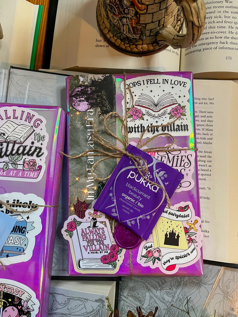 K&ouml;nnte beinhalten: Ein lila und pinkfarbenes Buch, das mit Schnur umwickelt ist und verschiedene Aufkleber hat, darunter "Always Falling for the Villain", "Oops I Fell in Love with the Villain" und "But they're spicier now". Ein lila Teebeutel mit dem Text "Pukka blackcurrant beauty organic/bio" ist ebenfalls sichtbar.