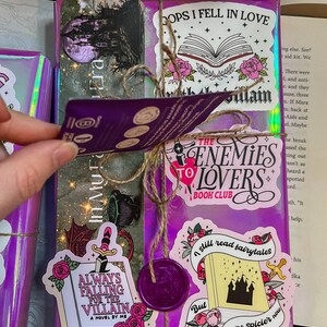 K&ouml;nnte beinhalten: Ein lilafarbenes Buch, das mit Schnur umwickelt ist und mit verschiedenen Aufklebern verziert ist. Die Aufkleber zeigen Illustrationen und Texte zum Thema B&uuml;cher, darunter "Always Falling for the Villain", "The Enemies to Lovers Book Club", "I still read fairytales, but they're spicier now" und "Oops I fell in love with the villain".