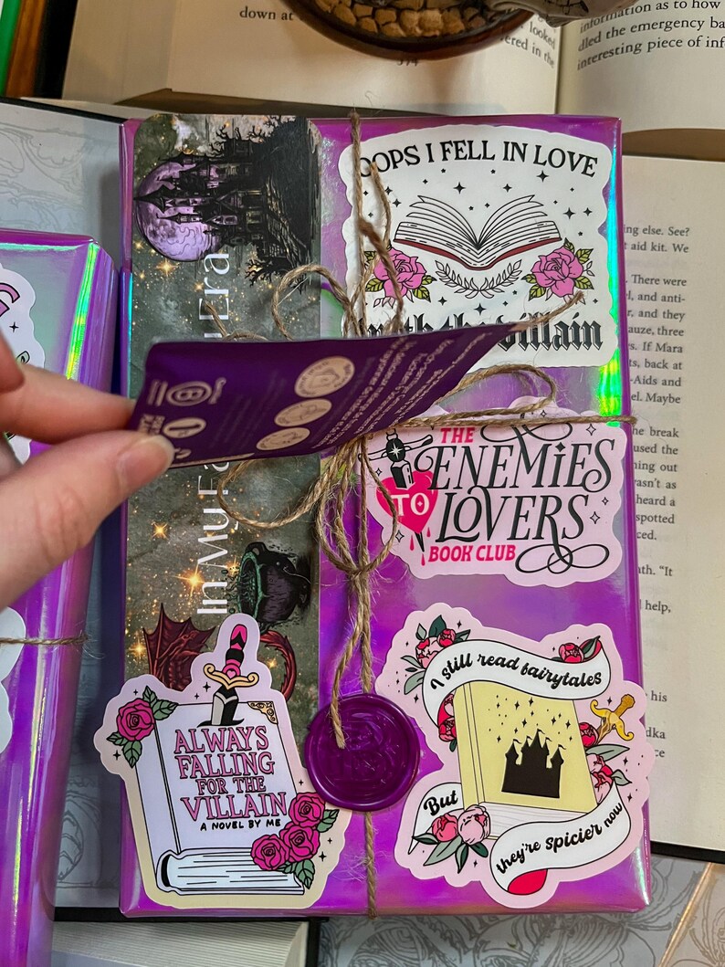 K&ouml;nnte beinhalten: Ein lilafarbenes Buch, das mit Schnur umwickelt ist und mit verschiedenen Aufklebern verziert ist. Die Aufkleber zeigen Illustrationen und Texte zum Thema B&uuml;cher, darunter "Always Falling for the Villain", "The Enemies to Lovers Book Club", "I still read fairytales, but they're spicier now" und "Oops I fell in love with the villain".