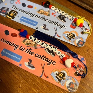 May include: Two decorative bookmarks with hockey-themed illustrations and the text "I'm coming to the cottage." One bookmark is light blue, the other is pink. Each has a beaded tassel with a name spelled out. Hockey jerseys, skates, and pucks are featured.