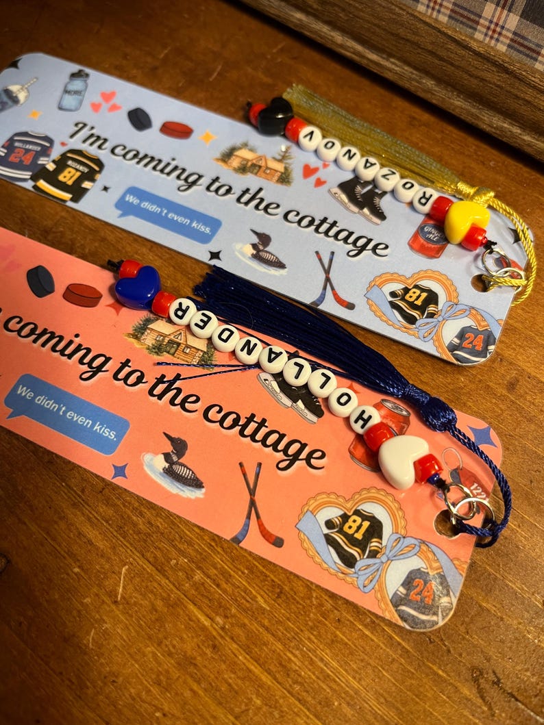 May include: Two decorative bookmarks with hockey-themed illustrations and the text "I'm coming to the cottage." One bookmark is light blue, the other is pink. Each has a beaded tassel with a name spelled out. Hockey jerseys, skates, and pucks are featured.