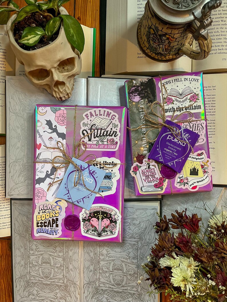 K&ouml;nnte beinhalten: Zwei verpackte B&uuml;cher mit bunten Aufklebern und einem Teebeutel. Die B&uuml;cher sind in schimmerndes, violettes Papier eingewickelt mit verschiedenen Aufklebern, darunter "Falling for the Villain", "Read Books Escape Reality" und "Always Falling for the Villain". Der Teebeutel ist mit schwarzer Johannisbeere aromatisiert und tr&auml;gt die Aufschrift "Pukka".