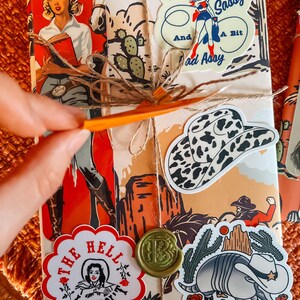 May include: A wrapped gift with a variety of colorful stickers. The stickers feature a cowgirl, a cactus, a armadillo, a hat, and a seal with the letter "B". The text on the stickers reads "Classy Sassy And A Bit Bad Ass", "The Hell I Won't", and "B".